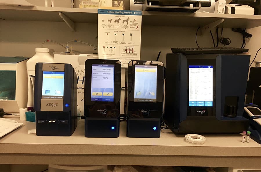 Three veterinary diagnostic analyzers are lined up on a lab counter, displaying various screens with sample data. Lab supplies and a sample handling chart are visible in the background.