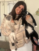 A woman with long brown hair smiles while holding a black-and-white cat in her right arm and a gray cat in her left arm, standing indoors near a window with blinds.