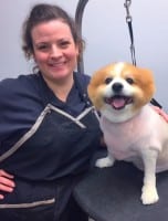 A woman in grooming attire stands next to a small dog with a fresh haircut, both facing the camera and smiling.