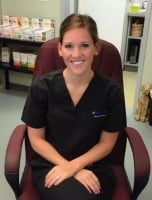 A woman in black scrubs sits in a maroon chair, smiling at the camera in a medical or veterinary office setting.