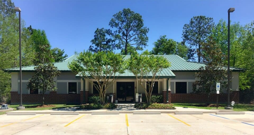 Single-story brick and stucco building with green roof, surrounded by trees, and an empty parking lot in front under clear blue sky.