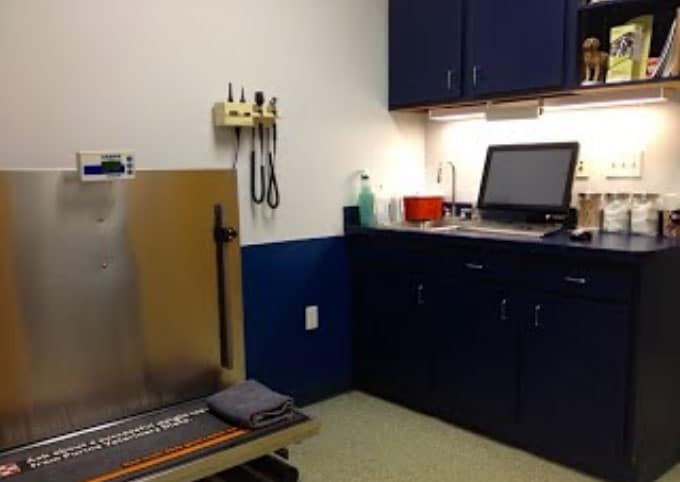 A veterinary exam room with a metal scale table, medical tools on the wall, a computer on a counter, and cabinets for storage.