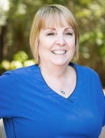 Smiling woman with short blonde hair wearing a blue scrub top, standing outdoors with greenery in the background.
