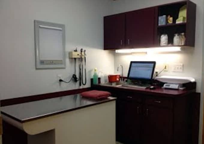 Medical exam room with an exam table, computer on a counter, medical equipment on the wall, and cabinets above and below the counter.