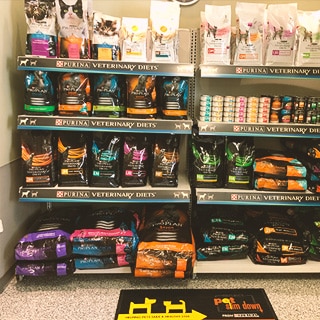 Shelves stocked with various bags of Purina Veterinary Diets pet food and cans in a pet store, with a yellow floor mat in front.