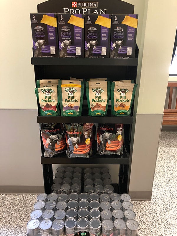 Store display with Purina Pro Plan dental chews on the top shelf, Greenies Pill Pockets in the middle, dog snacks below, and stacked cans of pet food on the bottom.