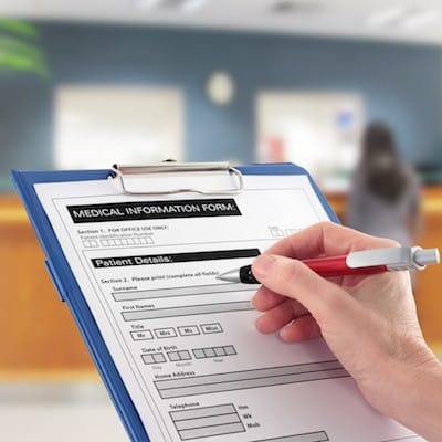 A person fills out a medical information form on a clipboard with a pen; a blurred hospital or clinic reception area is visible in the background.