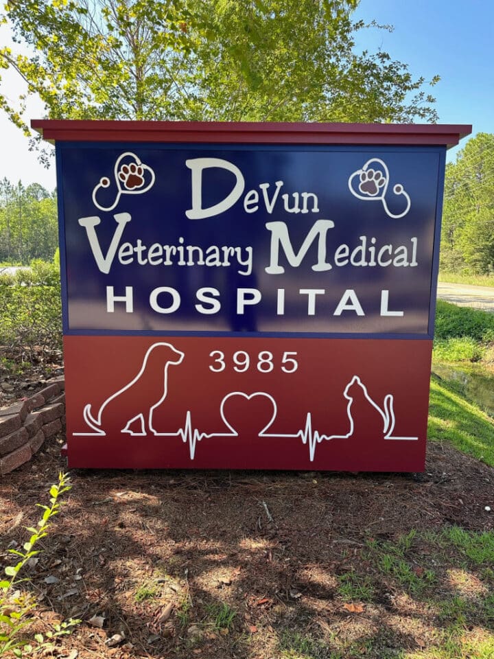 Outdoor sign for DeVun Veterinary Medical Hospital with dog, cat, heart, and paw print graphics. Street number 3985 is displayed. Trees and grass are visible in the background.