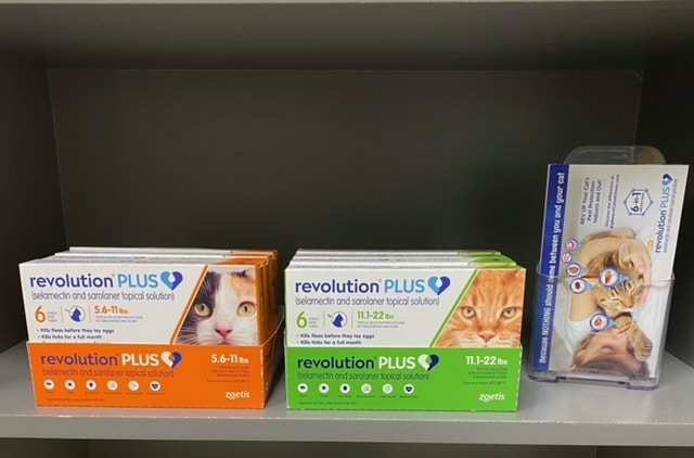 Revolution Plus feline medication boxes and a display of tick/flea preventatives are arranged on a grey shelf.