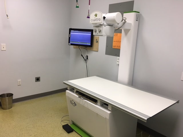 A veterinary X-ray machine with a flat examination table, wall-mounted display screen, and medical equipment in a clean, well-lit room.