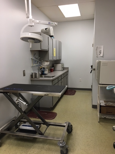 A medical examination room with an adjustable exam table, overhead light, cabinets, counter with medical supplies, and a small refrigerator.