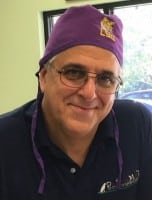 A man wearing glasses and a purple LSU cap smiles while seated indoors, with a window and greenery visible in the background.