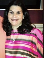 Woman with long dark hair wearing a pink, green, and white striped top, smiling at the camera in an indoor setting.