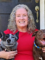 A woman with curly gray hair in a red shirt sits in front of a door, smiling, with two dogs—one black and white, one brown—on either side of her.