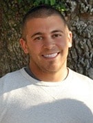 A person with short brown hair, a trimmed beard, and a light gray shirt is smiling while standing outdoors in front of a tree.