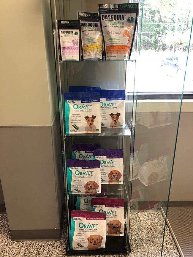 A glass display case holds packages of Dasuquin supplements on the top shelf and Oravet dental chews for dogs on the lower shelves, next to a window in a veterinary clinic.
