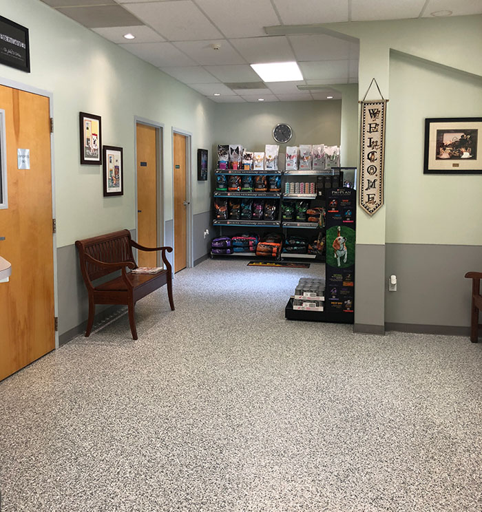 A clean, well-lit hallway with wooden doors, a bench, framed pictures, and shelves displaying pet food and supplies. A “Welcome” sign hangs on the wall.